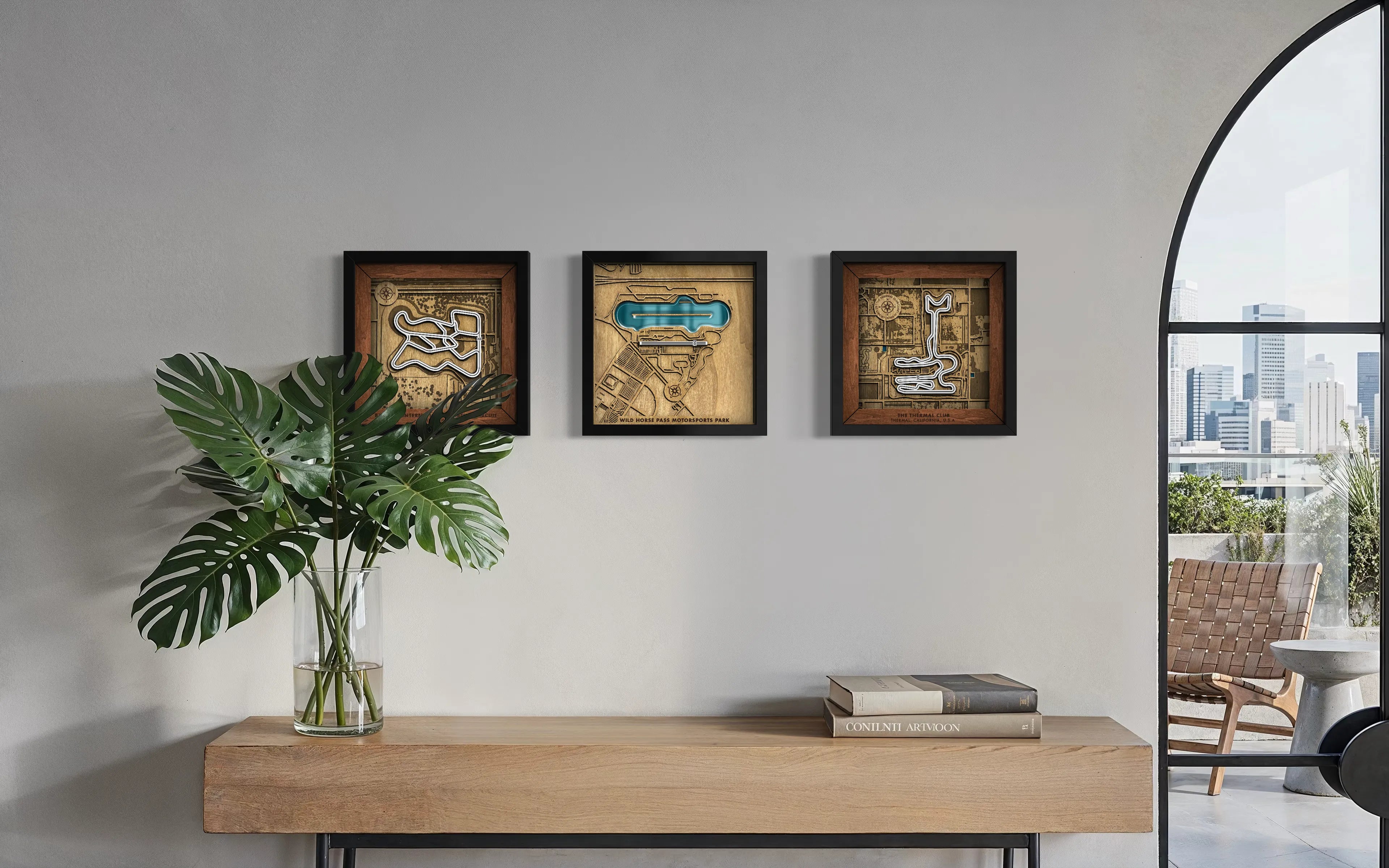 Three framed abstract art prints with black frames hang on a light gray wall above a wooden console table. A large green plant in a glass vase sits on the left, and two books rest on the right.
