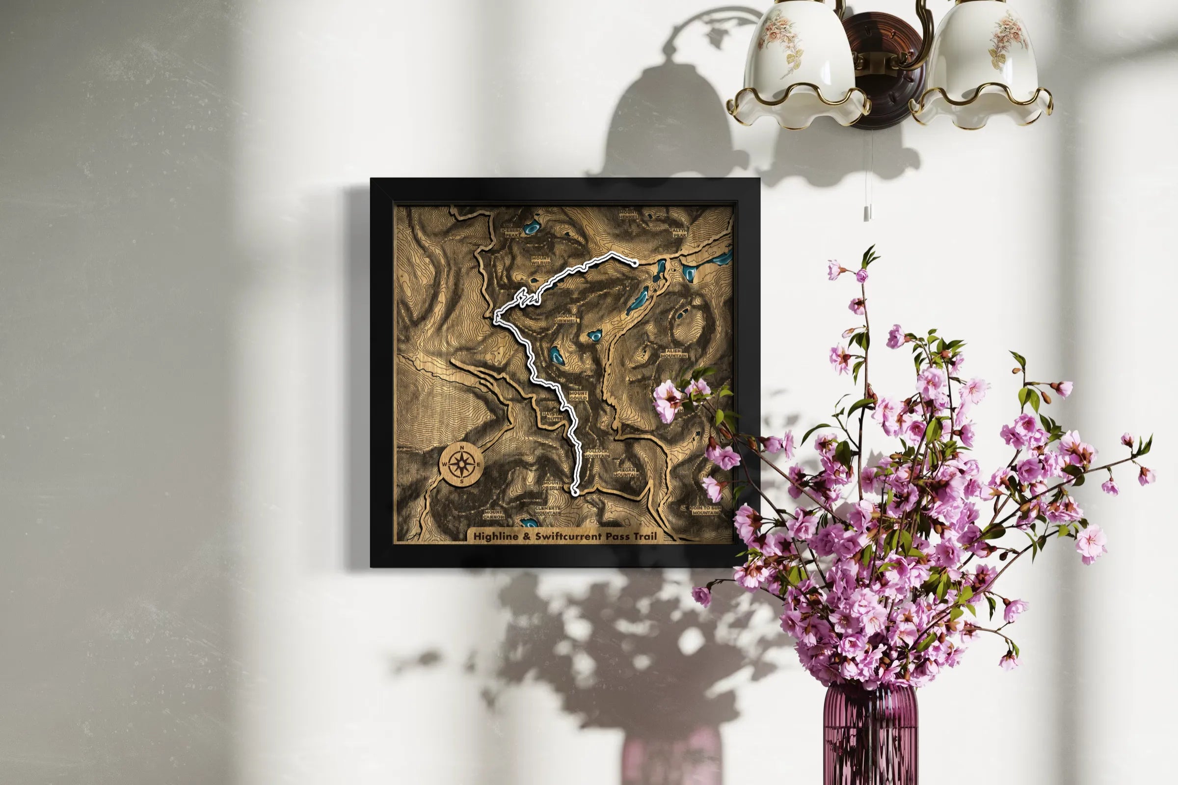 A framed 3D wooden topographic map art piece hanging on a white wall, styled with a pink flowering branch in a vase and a wall sconce casting shadows.
