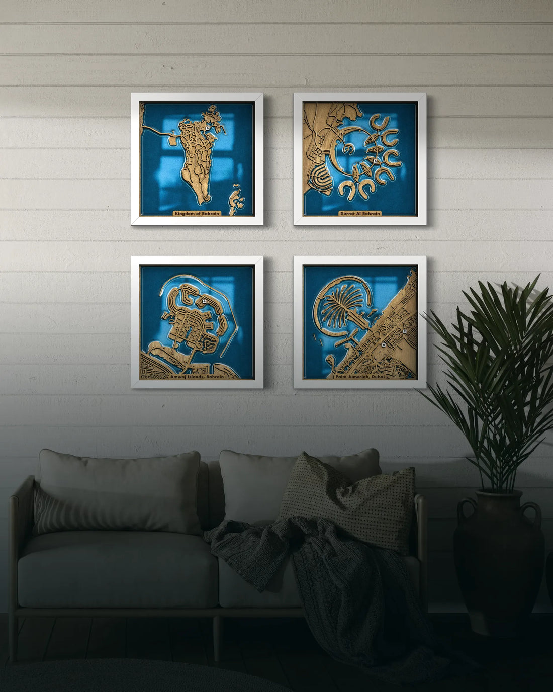 Four framed art prints with gold and blue designs are displayed on a white paneled wall above a gray sofa with pillows and a throw blanket, next to a potted plant.
