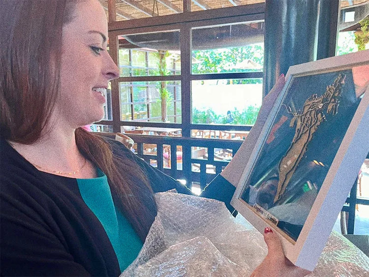 A woman holding a framed artwork inside what appears to be a rustic indoor gallery or shop, with large windows showing greenery outside.