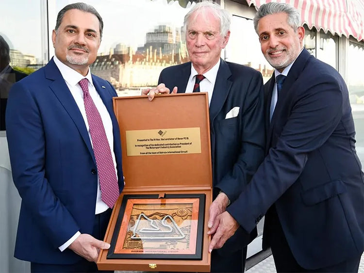 Three men in business suits stand together holding an open wooden presentation box containing a framed Masonic square and compass plaque. The box lid has a gold plaque, and the background shows an outdoor architectural setting with blurred buildings and a striped awning.