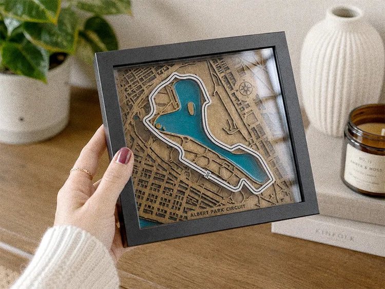 A hand holds a square framed 3D map art piece of Albert Park Circuit, featuring a brown wood-grain street map with a raised blue lake and white circuit track, in a cozy home setting.